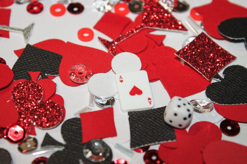 May include: A collection of red, black, and white casino-themed decorations. Includes playing card suit shapes, a die, an Ace of Hearts playing card, and sparkling embellishments. The items are scattered on a white surface.