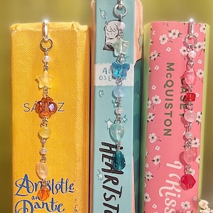 Op de afbeelding: Drie boeken met decoratieve kralen bladwijzers. De boeken heten "Aristotle and Dante Dive into the Waters of the World", "Heartstopper Volume 2" en "I Kissed Shara Wheeler". De bladwijzers hebben verschillende kralen en bedels.
