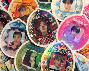 Puede incluir: Una colección de llaveros Tamagotchi holográficos y coloridos con diferentes imágenes de ídolos del K-pop. Los llaveros son redondos y tienen un diseño de pantalla digital con el texto "Tamagotchi" impreso en ellos.