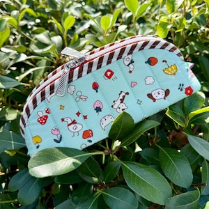 May include: A light blue quilted zippered pouch featuring a repeating pattern of small colorful illustrations including ducks, mushrooms, strawberries, flowers, and umbrellas, finished with a brown and white striped fabric trim along the top edge.