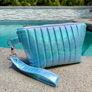 May include: A teal iridescent clutch with a vertical quilted design. The clutch has a matching wrist strap attached with a rose gold clasp. The zipper is silver.