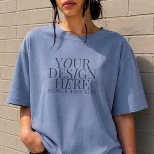 May include: A pale blue t-shirt featuring the text "YOUR DESIGN HERE" and "SNOWDROPMOCKUPS". The shirt has short sleeves and a loose fit. The model is wearing black jeans. The background is a brick wall.