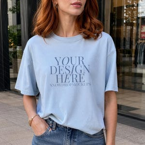 Peut inclure: Un t-shirt bleu clair avec le texte "YOUR DESIGN HERE" et "SNOWDROPMOCKUPS" en bleu plus foncé. Le modèle porte une jupe en jean et des bracelets dorés. L'arrière-plan est une vitrine de magasin.