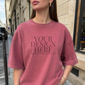 Peut inclure: Un t-shirt rose poussiéreux avec le texte "YOUR DESIGN HERE" et "SNOWDROPMOCKUPS" imprimé sur le devant. Le t-shirt a des manches courtes et une coupe décontractée. Le modèle porte un jean bleu.