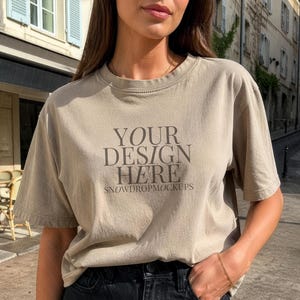 May include: A beige t-shirt with the text "YOUR DESIGN HERE" and "SNOWDROPMOCKUPS" printed on the front. The model is wearing black jeans. The background shows a European street scene.
