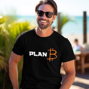 Könnte beinhalten: Schwarzes T-Shirt mit dem Wort "PLAN" in weißer, verwaschener Schrift und einem orangefarbenen Bitcoin-Symbol. Das T-Shirt hat kurze Ärmel.