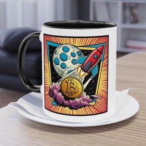 May include: White ceramic coffee mug with a black handle and interior. The mug features a colorful graphic of a rocket ship blasting off toward a moon, with a Bitcoin symbol in the foreground. The design is framed in a retro style.