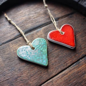 May include: Two ceramic heart ornaments hanging from twine. One heart is red and the other is teal with white speckles. The hearts are hanging on a rustic wooden surface.