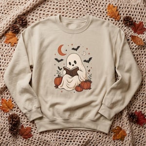 Peut inclure: Un sweat-shirt à col rond beige avec un motif central représentant un fantôme blanc lisant un livre, entouré de citrouilles orange, de chauves-souris noires, d'un croissant de lune et de petites étoiles dans des tons orange, marron et noir.