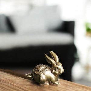 May include: A small, detailed brass rabbit figurine. The bunny is in a seated position with long ears and intricate patterns on its back. The figurine has a vintage look and is displayed on a wooden surface.