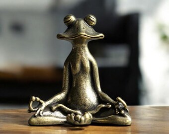 Könnte beinhalten: Eine goldfarbene Froschfigur aus Metall, die in meditativer Yoga-Pose sitzt. Der Frosch hat einen ruhigen Gesichtsausdruck.