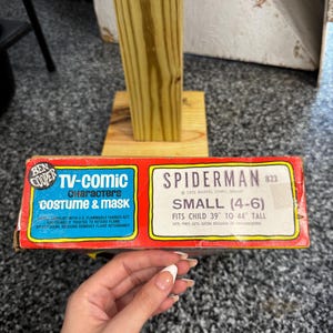 May include: A vintage Ben Cooper Spiderman TV-comic costume and mask box. The red and yellow packaging features blue text reading TV-Comic Characters Costume & Mask and Spiderman, indicating a size small for children 39 to 44 inches tall.