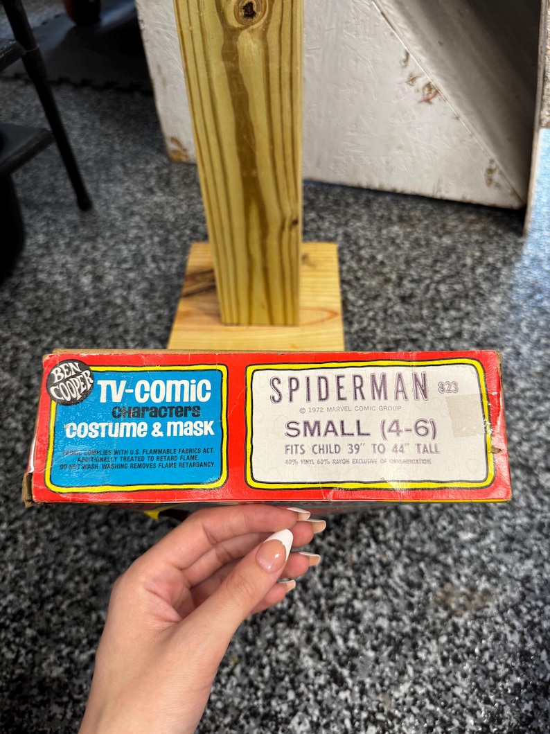 May include: A vintage Ben Cooper Spiderman TV-comic costume and mask box. The red and yellow packaging features blue text reading TV-Comic Characters Costume & Mask and Spiderman, indicating a size small for children 39 to 44 inches tall.