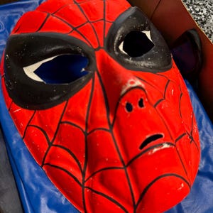 May include: A red plastic mask featuring a black web pattern across the surface. The mask has large, almond-shaped black eye cutouts with white borders, a molded nose bridge, and a small horizontal slit for the mouth area, showing a glossy finish.