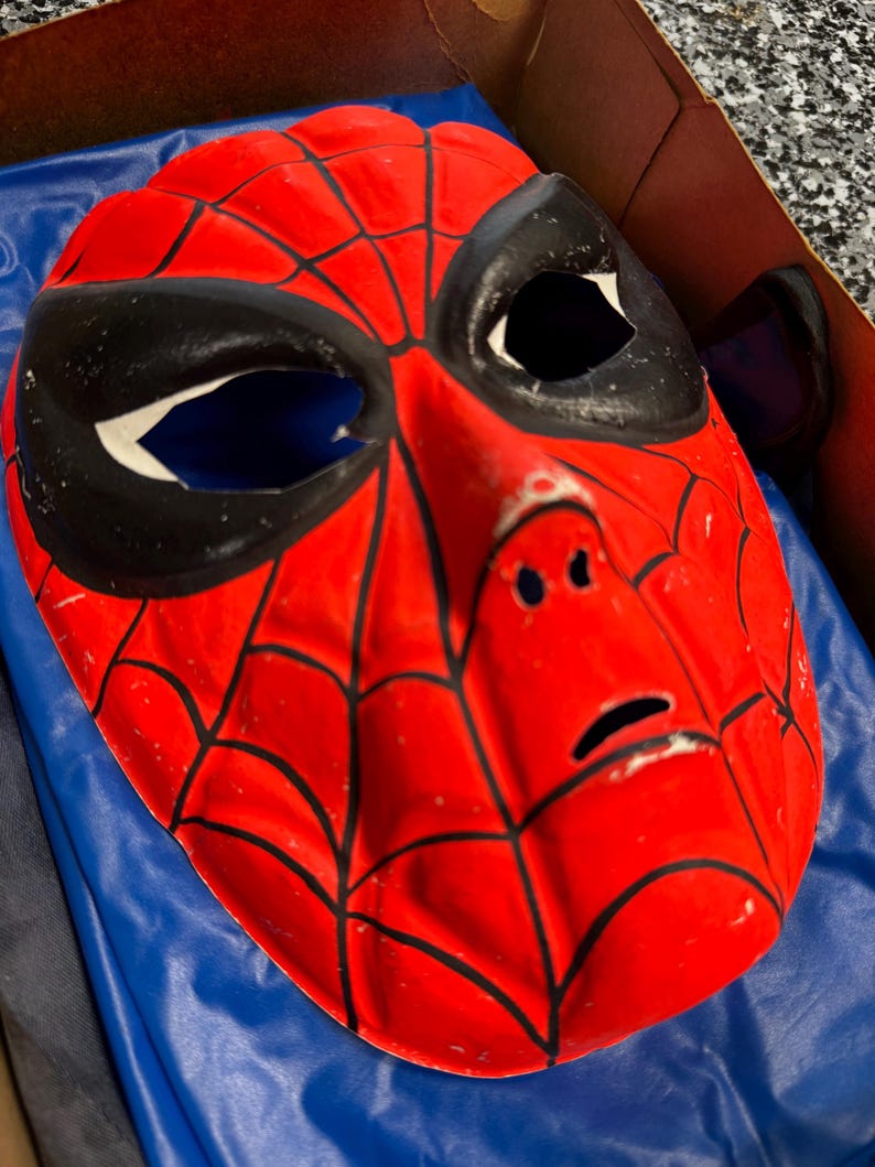 May include: A red plastic mask featuring a black web pattern across the surface. The mask has large, almond-shaped black eye cutouts with white borders, a molded nose bridge, and a small horizontal slit for the mouth area, showing a glossy finish.