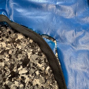 May include: A smooth, vibrant blue material with a slightly wrinkled texture, featuring a curved edge finished with a thick, black fabric binding. The blue surface has a subtle sheen and is positioned against a speckled black, white, and gray surface.