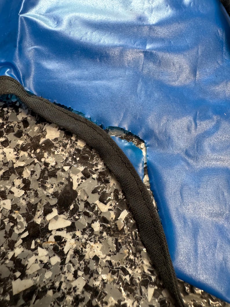 May include: A smooth, vibrant blue material with a slightly wrinkled texture, featuring a curved edge finished with a thick, black fabric binding. The blue surface has a subtle sheen and is positioned against a speckled black, white, and gray surface.
