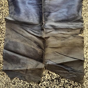 May include: A pair of fabric pants featuring a yellow and blue web pattern at the top with a red and black spider emblem. The lower portion is a solid dark blue fabric with visible stitching down the center and frayed edges on the right side.