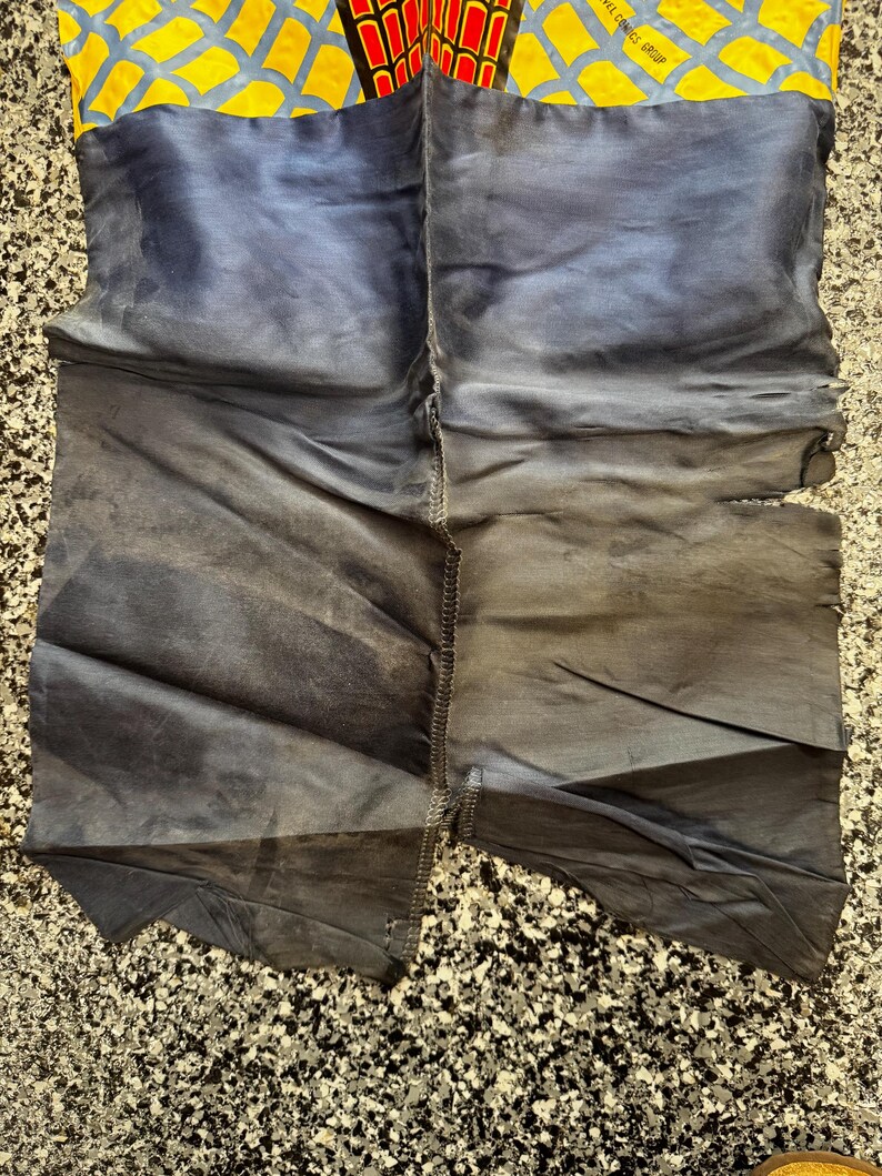 May include: A pair of fabric pants featuring a yellow and blue web pattern at the top with a red and black spider emblem. The lower portion is a solid dark blue fabric with visible stitching down the center and frayed edges on the right side.