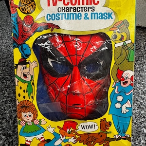 May include: A vintage Ben Cooper TV-Comic Characters costume and mask box featuring a red and black Spider-Man mask visible through a central cutout. The yellow packaging displays illustrations of various cartoon characters including Fred Flintstone.