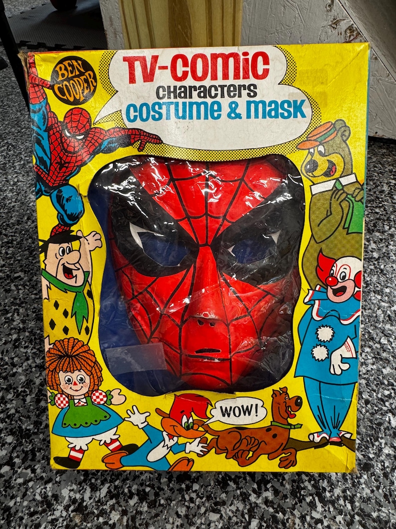 May include: A vintage Ben Cooper TV-Comic Characters costume and mask box featuring a red and black Spider-Man mask visible through a central cutout. The yellow packaging displays illustrations of various cartoon characters including Fred Flintstone.