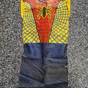 May include: A sleeveless Spider-Man costume featuring a red and yellow web pattern on the upper torso with a black spider emblem and the text Spider-Man. The lower portion is solid dark blue, and the garment includes blue ties at the neckline.