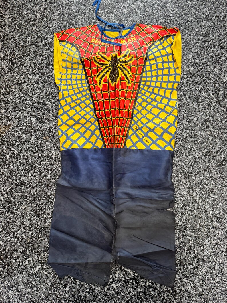 May include: A sleeveless Spider-Man costume featuring a red and yellow web pattern on the upper torso with a black spider emblem and the text Spider-Man. The lower portion is solid dark blue, and the garment includes blue ties at the neckline.