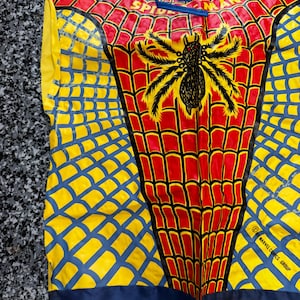 May include: A sleeveless costume top featuring a red and yellow spiderweb pattern with a large black spider graphic in the center. The text SPIDER-MAN is printed near the neckline, and the lower right side includes the text 2001 MARVEL COMICS GROUP.