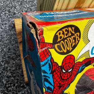 May include: A vintage Ben Cooper Spider-Man TV-comic costume and mask cardboard box featuring a colorful graphic of the character in red and blue on a yellow background, showing significant wear and a torn corner on the upper left side.