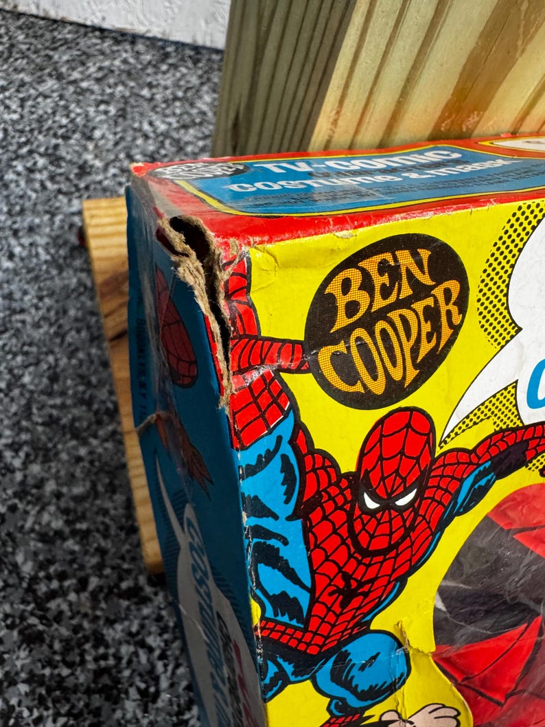 May include: A vintage Ben Cooper Spider-Man TV-comic costume and mask cardboard box featuring a colorful graphic of the character in red and blue on a yellow background, showing significant wear and a torn corner on the upper left side.