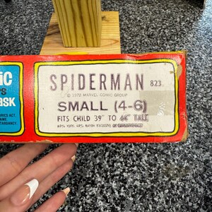May include: A rectangular red cardboard box for a Spiderman mask, model 823, dated 1972 Marvel Comic Group. The label specifies size small (4-6) for a child 39 inches to 44 inches tall, noting 40% vinyl and 60% rayon material composition.