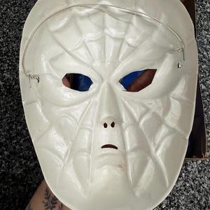 May include: A white plastic mask featuring a molded spiderweb pattern across the entire surface. The mask includes cutouts for the eyes, nose, and mouth, and is secured with a thin, flexible silver-colored wire strap attached to the sides.