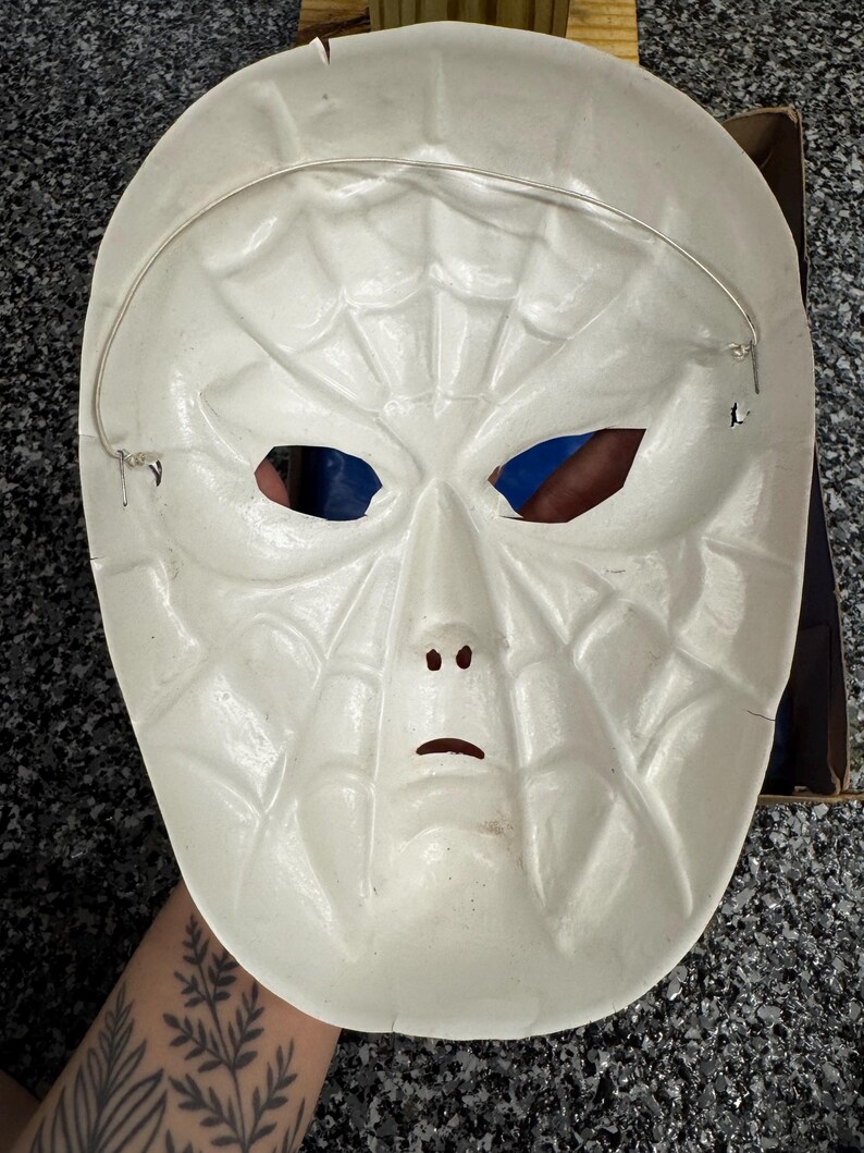 May include: A white plastic mask featuring a molded spiderweb pattern across the entire surface. The mask includes cutouts for the eyes, nose, and mouth, and is secured with a thin, flexible silver-colored wire strap attached to the sides.