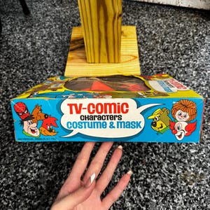May include: A vintage Ben Cooper TV-Comic Characters costume and mask box featuring colorful illustrations of various cartoon characters including Spider-Man, Yogi Bear, and a clown. The blue and yellow box has a clear plastic window on top.