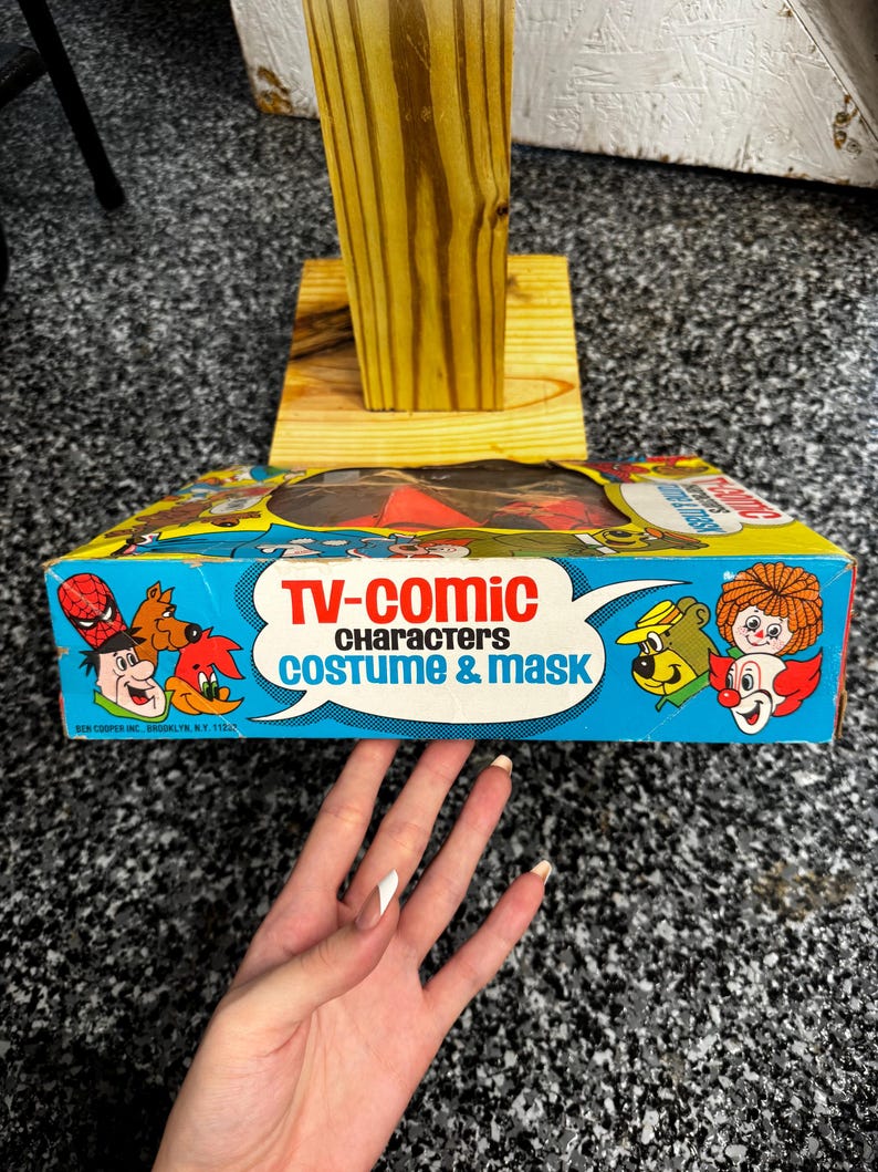 May include: A vintage Ben Cooper TV-Comic Characters costume and mask box featuring colorful illustrations of various cartoon characters including Spider-Man, Yogi Bear, and a clown. The blue and yellow box has a clear plastic window on top.