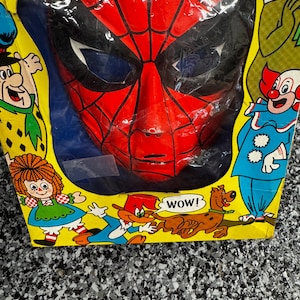 May include: A red and black Spider-Man mask encased in a yellow cardboard box. The packaging features colorful illustrations of various cartoon characters, including a clown, a dog, and a girl with red hair, with the text WOW! printed near the dog.