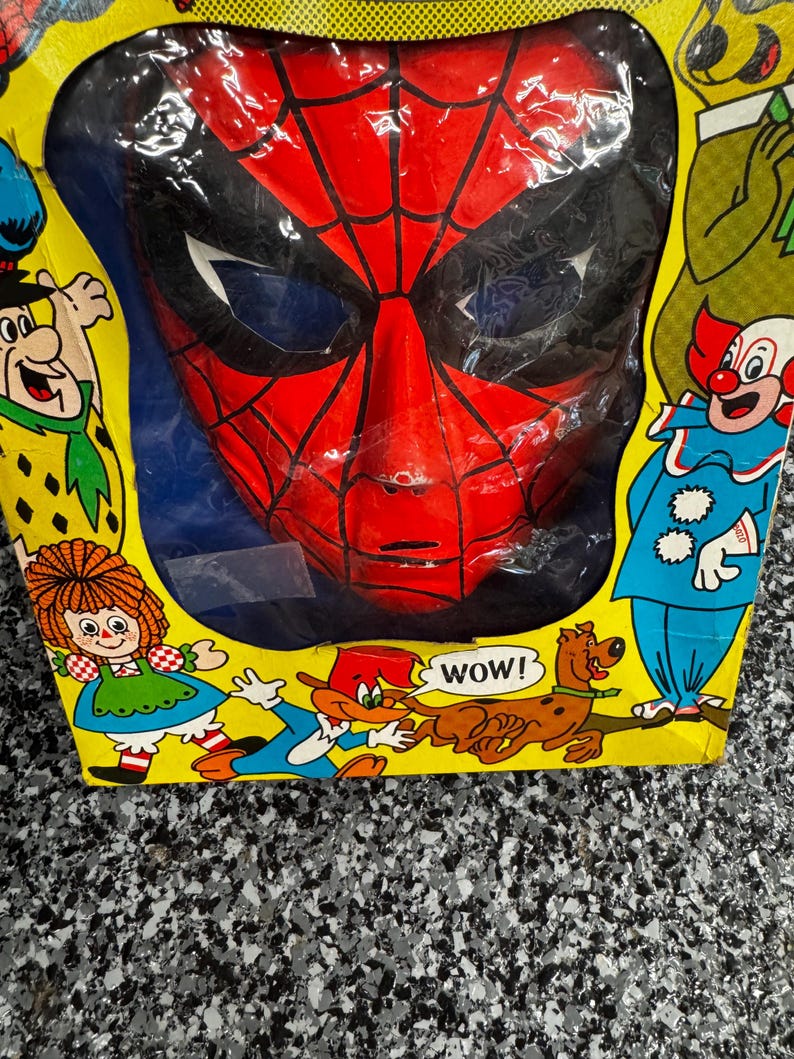 May include: A red and black Spider-Man mask encased in a yellow cardboard box. The packaging features colorful illustrations of various cartoon characters, including a clown, a dog, and a girl with red hair, with the text WOW! printed near the dog.