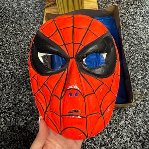 May include: A bright red plastic mask featuring a black spiderweb pattern across the surface. The mask has large, almond-shaped black eye cutouts, a molded nose bridge, and a small horizontal slit for the mouth area, with visible paint wear.