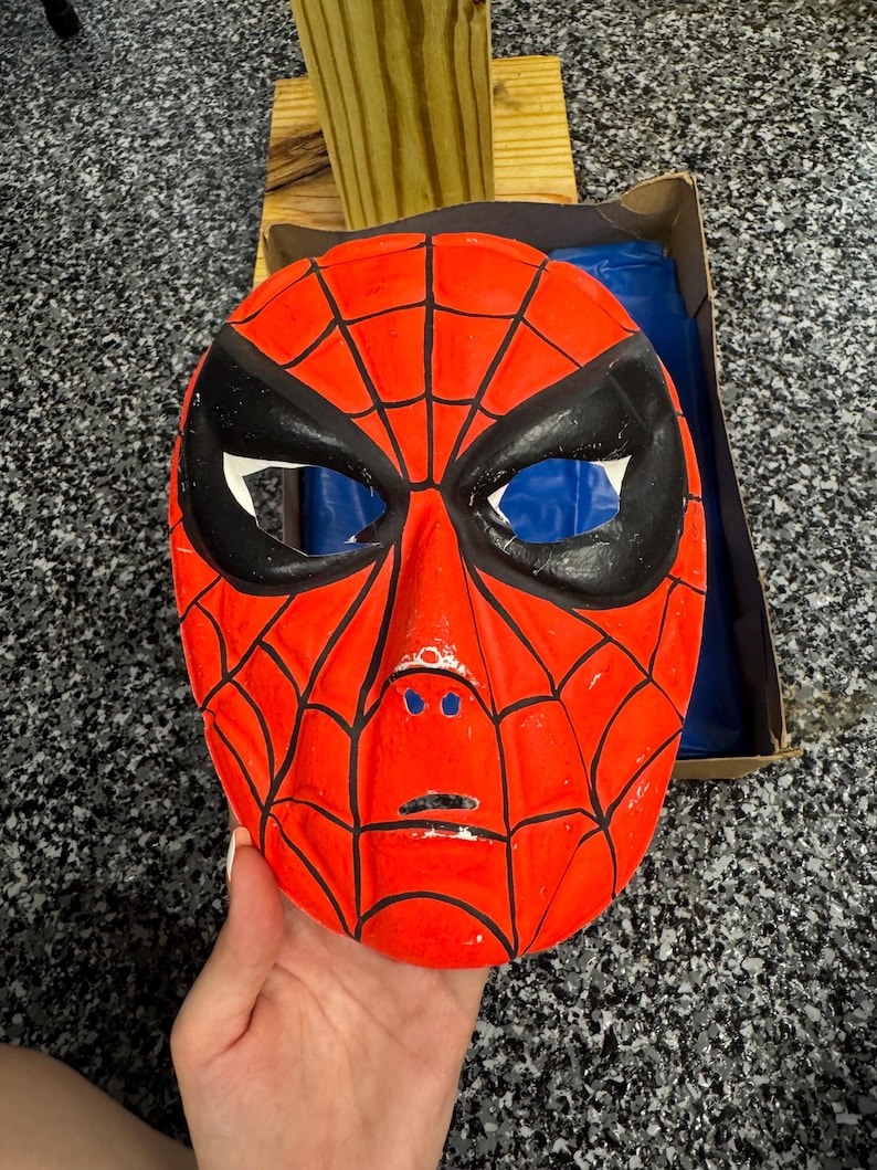 May include: A bright red plastic mask featuring a black spiderweb pattern across the surface. The mask has large, almond-shaped black eye cutouts, a molded nose bridge, and a small horizontal slit for the mouth area, with visible paint wear.
