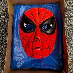 May include: A red and black Spider-Man mask with a web pattern and large eye openings, resting on a blue plastic sheet inside a brown cardboard box. The mask features a molded face shape with black outlines around the eyes and across the surface.