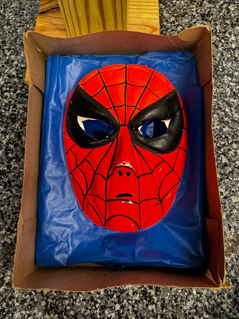 May include: A red and black Spider-Man mask with a web pattern and large eye openings, resting on a blue plastic sheet inside a brown cardboard box. The mask features a molded face shape with black outlines around the eyes and across the surface.