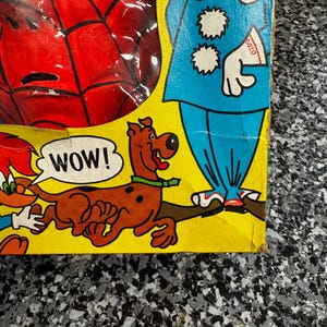 May include: A yellow cardboard toy box featuring a red Spider-Man mask visible through a plastic window, a blue clown illustration, and a brown Scooby-Doo character with a speech bubble containing the text WOW! printed in black capital letters.