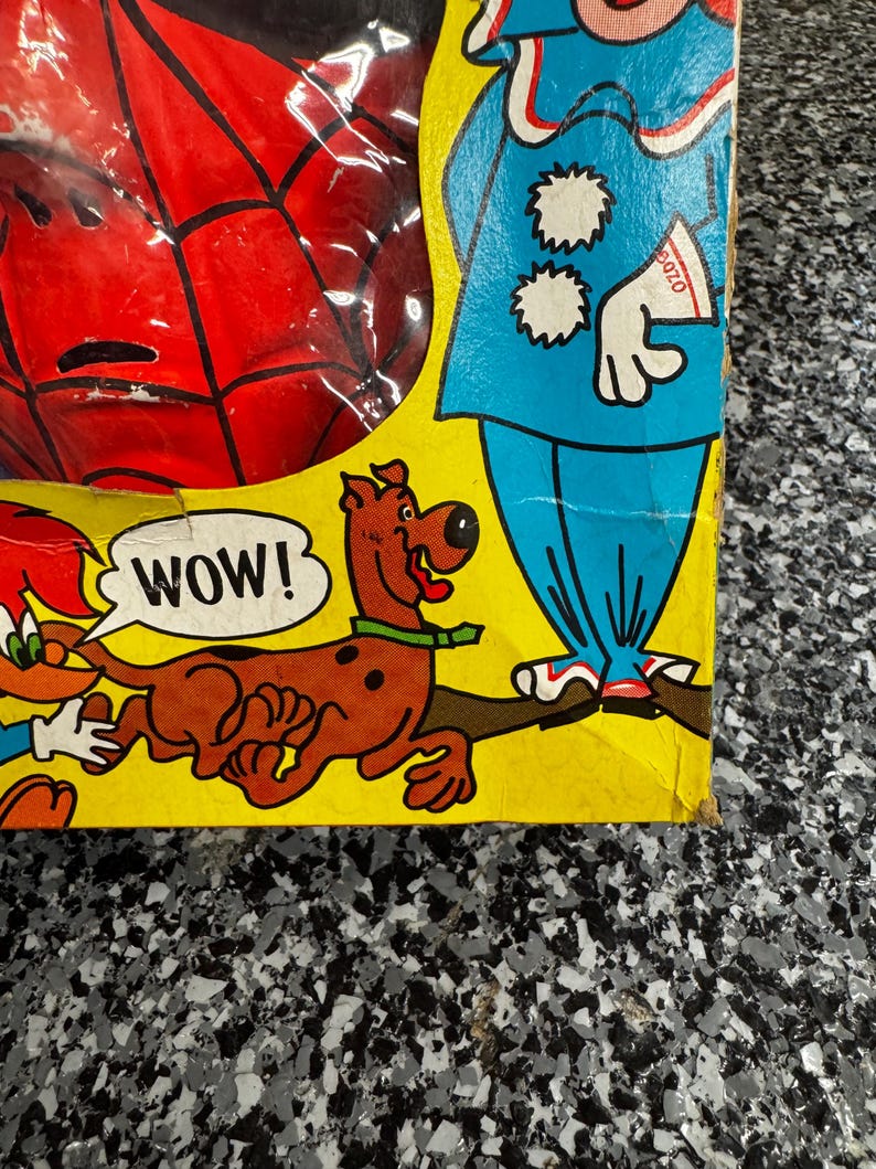 May include: A yellow cardboard toy box featuring a red Spider-Man mask visible through a plastic window, a blue clown illustration, and a brown Scooby-Doo character with a speech bubble containing the text WOW! printed in black capital letters.