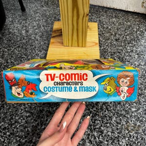 May include: A rectangular cardboard box for a TV-Comic Characters Costume & Mask set. The blue packaging features colorful illustrations of various cartoon characters, including a clown, a bear, and a bird, with bold red and blue text.