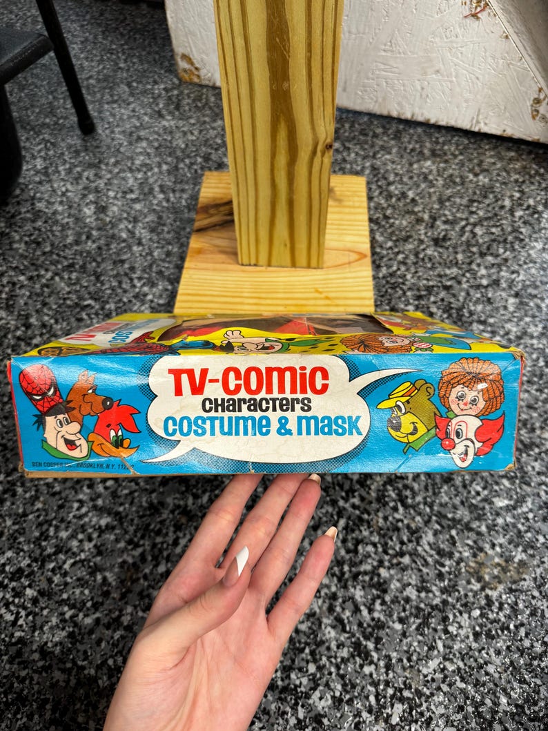 May include: A rectangular cardboard box for a TV-Comic Characters Costume & Mask set. The blue packaging features colorful illustrations of various cartoon characters, including a clown, a bear, and a bird, with bold red and blue text.
