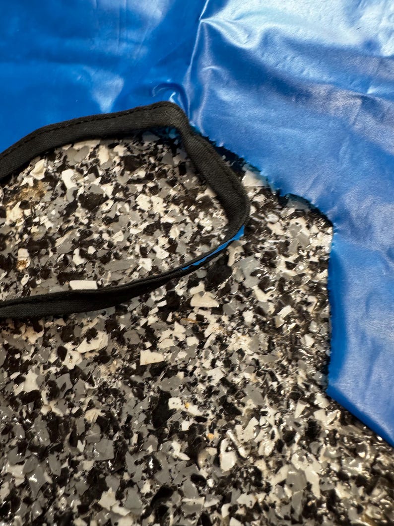 May include: A bright blue fabric piece with a smooth, slightly reflective surface and a jagged, uneven edge. The fabric features a black fabric trim stitched along a curved opening, creating a distinct contrast against the vibrant blue material.