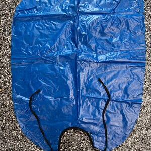 May include: A bright blue, rectangular plastic apron with a smooth, crinkled texture. The apron features a rounded neckline with thin, black fabric ties attached to the sides for securing the garment around the torso during use.