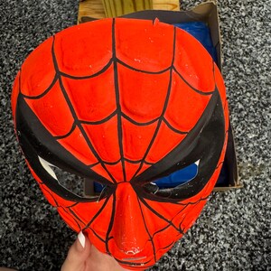 May include: A bright red plastic mask featuring a black web pattern across the surface. The mask has two large, angular black eye openings with white mesh inserts. The design mimics a comic book character, with a molded nose and chin area.