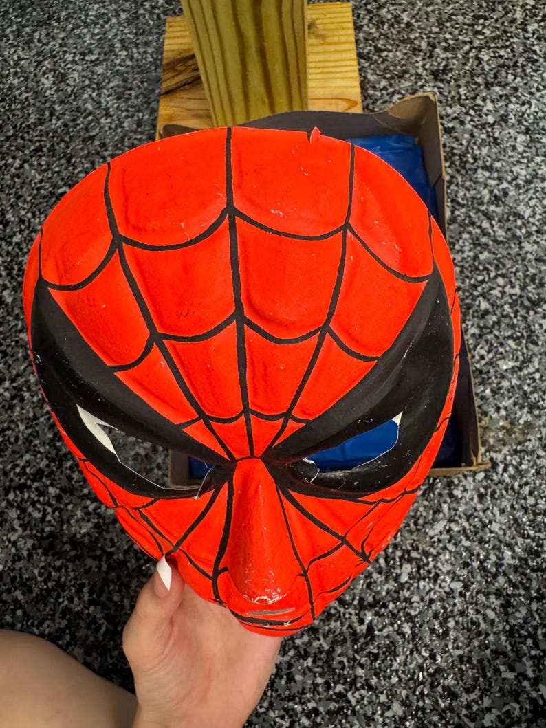 May include: A bright red plastic mask featuring a black web pattern across the surface. The mask has two large, angular black eye openings with white mesh inserts. The design mimics a comic book character, with a molded nose and chin area.