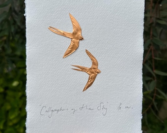 May include: A handmade artwork featuring two copper-colored swallows in flight, mounted on textured white paper. The artwork is titled "Calligraphers of The Sky" and measures ⅛ in.
